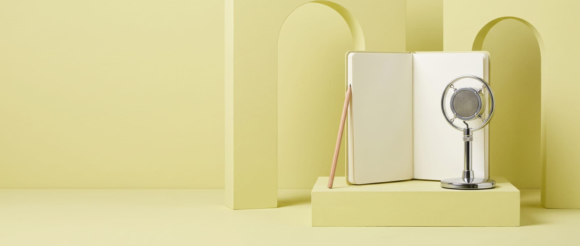 An open notebook and a chrome studio microphone on a pale yellow plinth